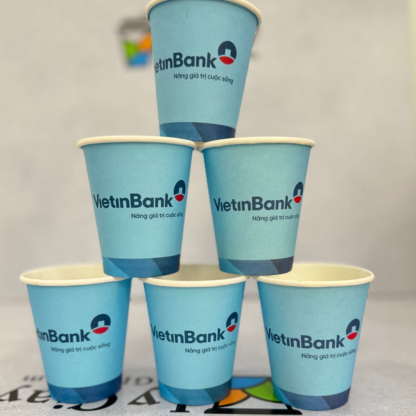 7 oz single wall paper cups printed pale blue with a bank logo, stacked in a pyramid